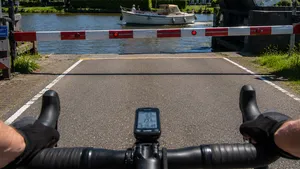 Een zwetende wielrenner met helm wacht in de volle zon voor een openstaande brug terwijl een traag bootje voorbijvaart.