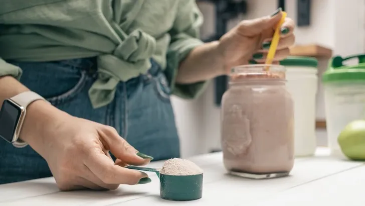 Young woman in jeans and shirt holding measuring spoon with protein powder, glass jar of protein drink cocktail, milkshake or smoothie above white wooden table with chocolate pieces, bananas, apples