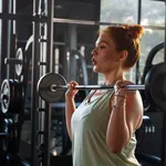 Focused woman performing barbell curls at gym, bathed in natural light. Gym workout routine.