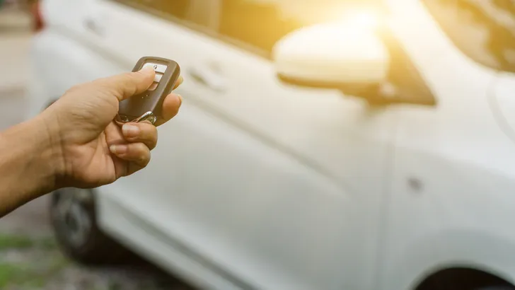 Hand richt een autosleutel op een witte auto en houdt een knop ingedrukt.