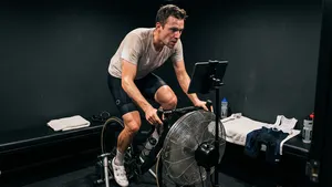 Een man op een indoor fietstrainer in een donkere kamer, intensief aan het zweten terwijl hij fietst. Een grote ventilator staat voor hem en een tablet is gemonteerd op een standaard. Op een bankje ernaast liggen handdoeken en een bidon.