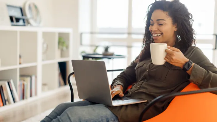 Smiling black lady watching video on computer, drinking hot coffee