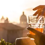 Young woman tourist fashion white dress with spritz cocktail in front of panoramic view of Rome cityscape from campidoglio terrace at sunset. Landmarks, domes of Rome, Italy.