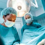 Closeup shot of young adult dedicated black female surgeon concentrated on performing operation standing under the surgical light with female colleague professionally dressed in operating gowns.