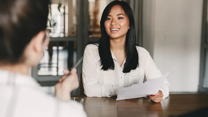 Business, career and placement concept - joyful asian woman smiling and holding resume, while sitting in front of directors during corporate meeting or job interview