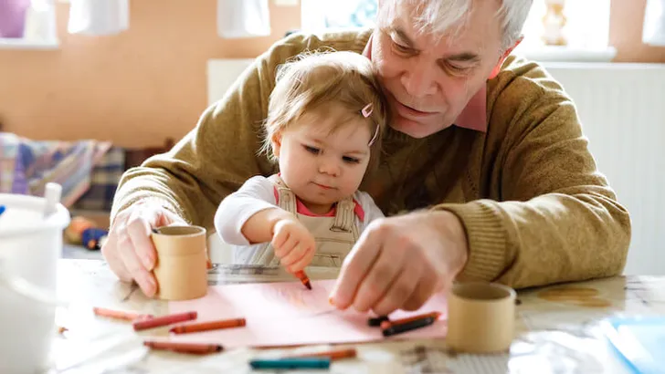 Cute little baby toddler girl and handsome senior grandfather painting with colorful pencils at home. Grandchild and man having fun together