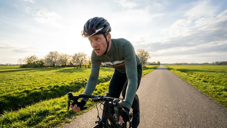 Mannelijke wielrenner met helm kijkt vermoeid maar voldaan tijdens zijn eerste rit in de lentezon over een polderweg, passend bij een wielrenplan opbouwen.