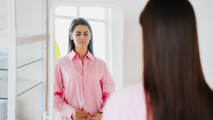Sad brunette fit young woman in pink oversize shirt stands in front of mirror feels loneliness. Frustrated hispanic girl in troubles standing on kitchen. Diet and health care. Dietetics. Fasting