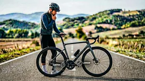 Een mannelijke wielrenner met helm kijkt nadenkend naar het crankstel van zijn racefiets, met een grafische overlay die het dilemma tussen een kortere en langere cranklengte symboliseert.