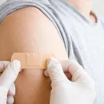 Doctor in white rubber protective gloves putting adhesive bandage on young man's arm after scratch on skin or injection of vaccine. First aid. Medical, pharmacy and healthcare concept. Closeup.