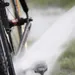 Een wielrenfiets wordt schoongespoten met een hogedrukspuit; waterstraal raakt trapas en frame