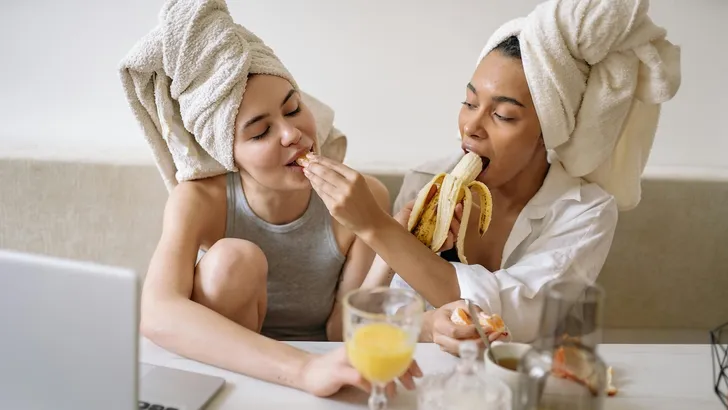 Experts waarschuwen voor beauty-hack met bananenschillen – Foto van twee vrouwen op een bank met haren in een handdoek gewikkeld en een banaan in de hand.