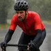 Wielrenner in felrode gabba rijdt door zware regen op natte weg, druppels parelen op het winddichte shirt en armstukken zijn zichtbaar.