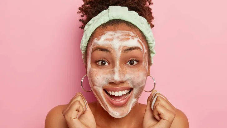 Close up shot of happy impressed curly African American woman raises clenched fists, gets pleasure from hygienic treatments, wears headband on head, washes face with bubble soap, smiles broadly