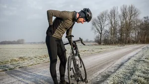 Een mannelijke wielrenner in volledige outfit met helm, die na zijn fietstocht met zijn hand zijn pijnlijke onderrug vastpakt, staand naast zijn racefiets in een Nederlands landschap.