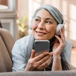 Happy caucasian mature woman listening to the music on the phone on the couch