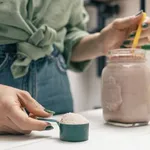 Young woman in jeans and shirt holding measuring spoon with protein powder, glass jar of protein drink cocktail, milkshake or smoothie above white wooden table with chocolate pieces, bananas, apples