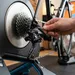Gedetailleerde close-up van een achterderailleur en cassette van een racefiets op een fietstrainer, waarbij een hand de stelschroef aanpast om de versnellingen af te stellen.