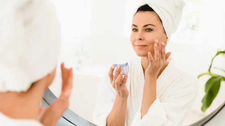 Caucasian middle-aged woman in turban and spa bathrobe looking at the mirror while applying beauty creme moisturizer for anti-age anti-wrinkle effect