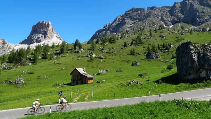 Twee wielrenners fietsen met snelheid over de asfaltweg van de Passo Gardena in de Italiaanse Dolomieten, omringd door groene alpenweides en de iconische rotsformaties onder een strakblauwe hemel.