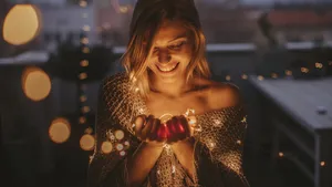 Young, smiling woman standing outdoors on the decorated terrace of her apartment and holding string lights