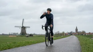 Mannelijke wielrenner met helm drinkt uit bidon op een Nederlandse polderweg.