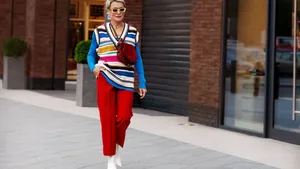 Fashionable Woman wearing trendy outfit, red trousers, blue jacket, striped vest, white shoes and yellow sunglasses. Model with short hair walking the street