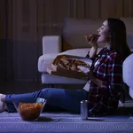 Young woman eating pizza while watching TV in room at night. Bad habit