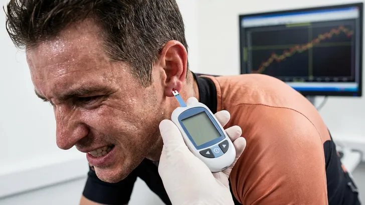 Een professionele wielrenner in een sportwetenschappelijk laboratorium, diep in inspanning op een fietstrainer met meetapparatuur en omringd door schermen vol prestatiedata en grafieken.