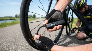 Een close-up van de handen van een mannelijke wielrenner met helm die geknield op het asfalt een moderne elektrische mini-compressor aansluit op het wiel van zijn racefiets.