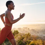 Running, black woman and fitness on sunset mountains, city background and blue sky for exercise goals, energy and power. Happy athlete, cardio and outdoor workout with motivation, smile and wellness