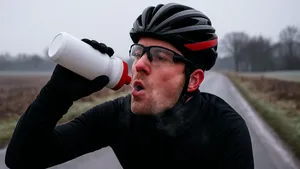 Close-up van een wielrenner in winterkleding die op een koude, mistige weg uit zijn bidon drinkt; ademwolk zichtbaar