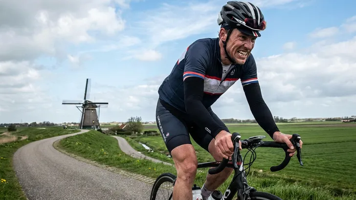 Een mannelijke wielrenner met helm en armstukken die zichtbaar afziet tijdens een zware inspanning op een dijk, met een molen in een typisch Nederlands polderlandschap op de achtergrond.