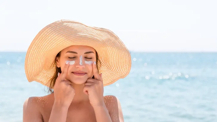 Beautiful young woman in hat is applying sunblock under her eyes and on her nose like Indian. Sun protection concept