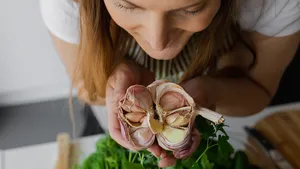 Close-up van een lachend vrouwengezicht terwijl ze een bol knoflook ruikt die op een keukentafel ligt.