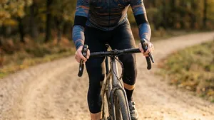 Een liggende foto van een mannelijke gravelbiker, gefotografeerd van de zijkant, gepositioneerd in het rechterderde deel van het frame, langs een slingerend grindpad door een herfstbos, met een handpositie op de gravelbike