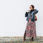 Smiling female dressed boho fashion style colorful long dress with black leather biker jacket with brown leather flap bag posing on the white wall background.