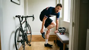 en wielrenner zit na zijn fietstocht op de grond en pakt met een pijnlijke uitdrukking de buitenkant van zijn voet vast, terwijl zijn racefiets tegen de muur staat.
