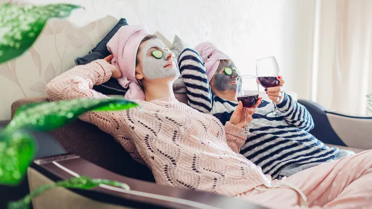 Mother and her adult daughter applied facial masks and cucumbers on eyes. Women chilling while having wine