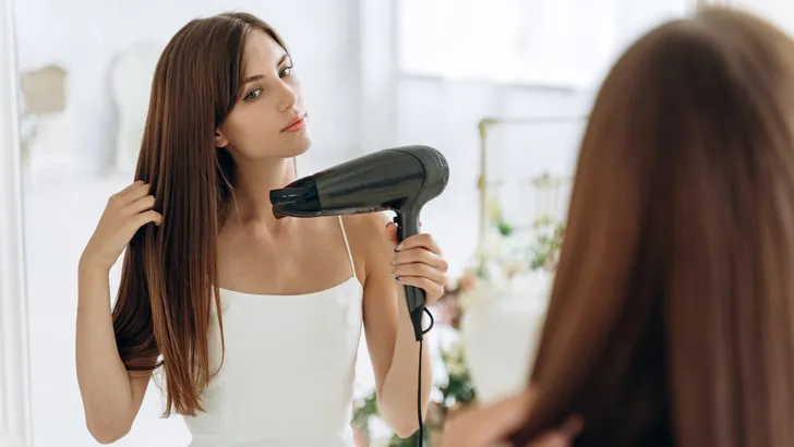 Experts delen tips om haargroei te helpen – Foto van vrouw met lang haar bij spiegel met föhn.