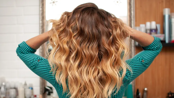 Dit kapsel zorgt voor extra volume bij fijn haar. – Foto van vrouw met lang golvend haar voor spiegel.