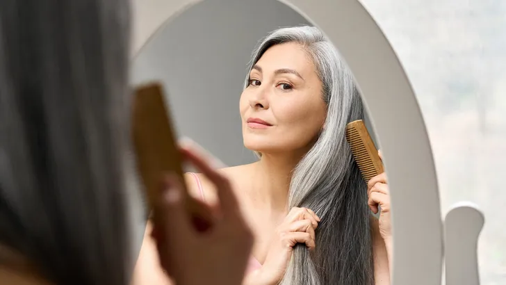 Mature mid aged senior Asian woman looking at mirror combing her gray hair.