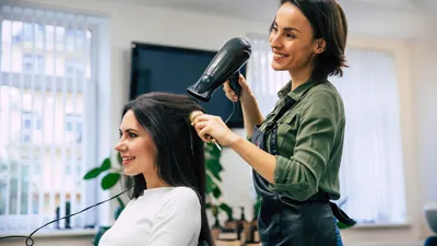 Close-up van een blije, lachende professionele styliste die het haar van haar tevreden klant föhnt en kamt.