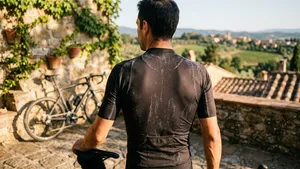 Een close-up van de rug van een mannelijke fietser die op een zonnig terras naast zijn fiets staat. Op zijn donkere fietsshirt zijn zoutvlekken van het zweet te zien, die oplichten in het warme zonlicht.