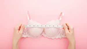 Young adult woman hands holding white measure tape and measuring bra on light pink table background. Pastel color. Closeup. Point of view shot. Choosing right size. Top down view.