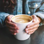 Closeup image of a woman holding a cup of hot latte coffee on the table