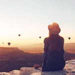 View from the back of a girl in a hat sits on a hill and looks at air balloons.