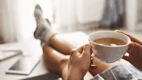Cup of tea and chill. Woman lying on couch, holding legs on coffee table, drinking hot coffee and enjoying morning, being in dreamy and relaxed mood. Girl in oversized shirt takes break at home