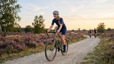 Een jonge Nederlandse vrouw fietst lachend op een matte gravelbike over een onverhard pad door de paarse heide bij zonsondergang, met een silhouet van een windmill op de achtergrond.