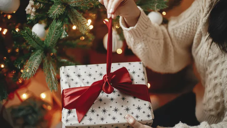 Hands in cozy sweater opening christmas gift with red bow on background of christmas tree with lights. Stylish female holding present with red ribbon in festive room close up. Merry Christmas!